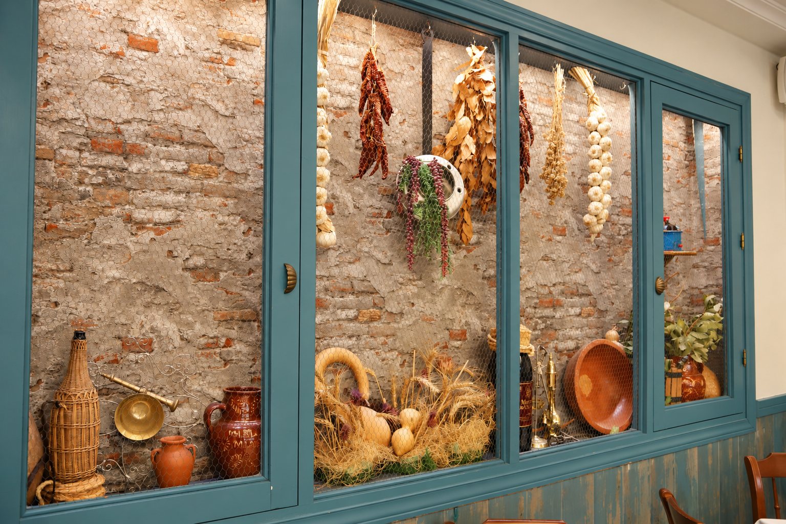 Ventana de carpintería azul con ajos, pimientos, espigas y cerámica tras el cristal, junto a las mesas.